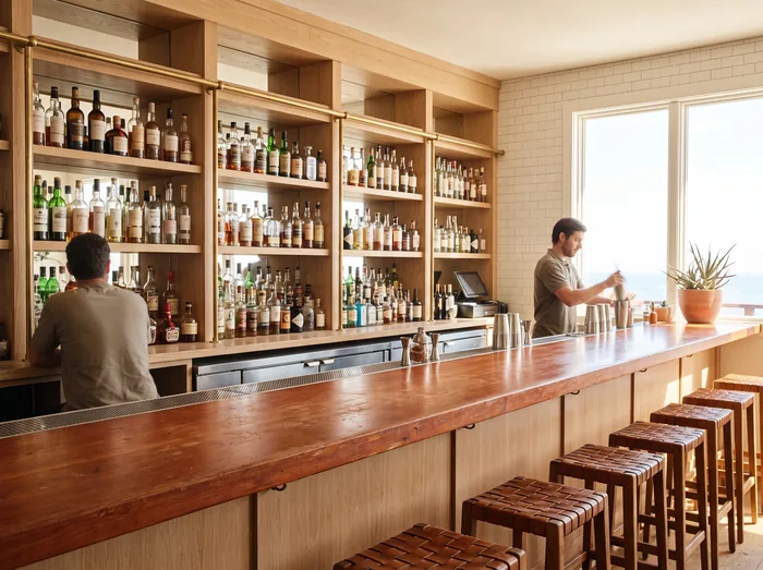 Interior of a bright coastal bar, a bartender mixing a drink in front of pale oak shelving stacked with bottles, ocean beyond the window.